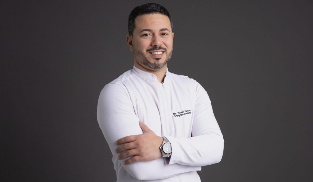 Fotografia profissional de um dentista sorridente usando jaleco branco, em frente a um fundo cinza escuro. Ele está com os braços cruzados, demonstrando confiança e simpatia. No jaleco, está bordado o nome “Dr. Paulo Ramos – Cirurgião-Dentista”. A imagem transmite profissionalismo e acolhimento, ideal para sites e materiais de clínicas odontológicas.