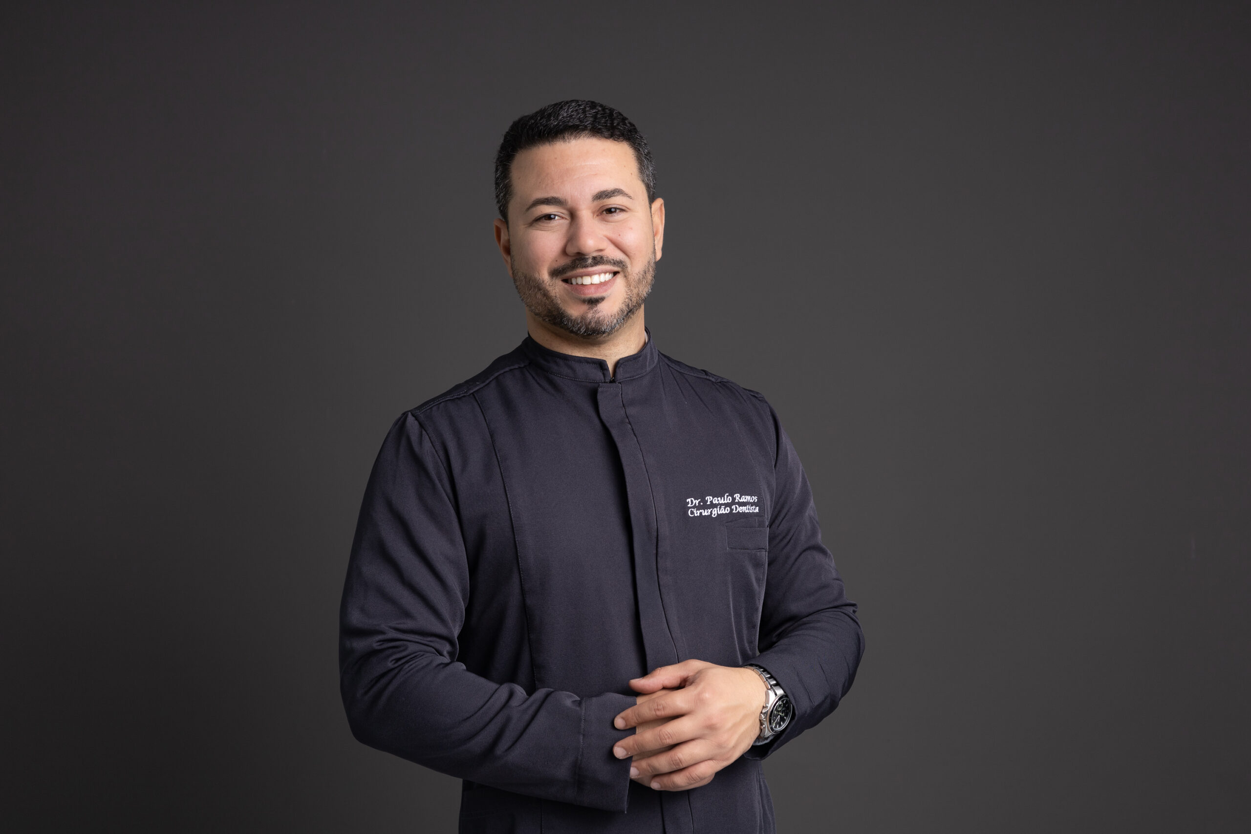 Fotografia profissional de um dentista sorridente usando jaleco branco, em frente a um fundo cinza escuro. Ele está com os braços cruzados, demonstrando confiança e simpatia. No jaleco, está bordado o nome “Dr. Paulo Ramos – Cirurgião-Dentista”. A imagem transmite profissionalismo e acolhimento, ideal para sites e materiais de clínicas odontológicas.