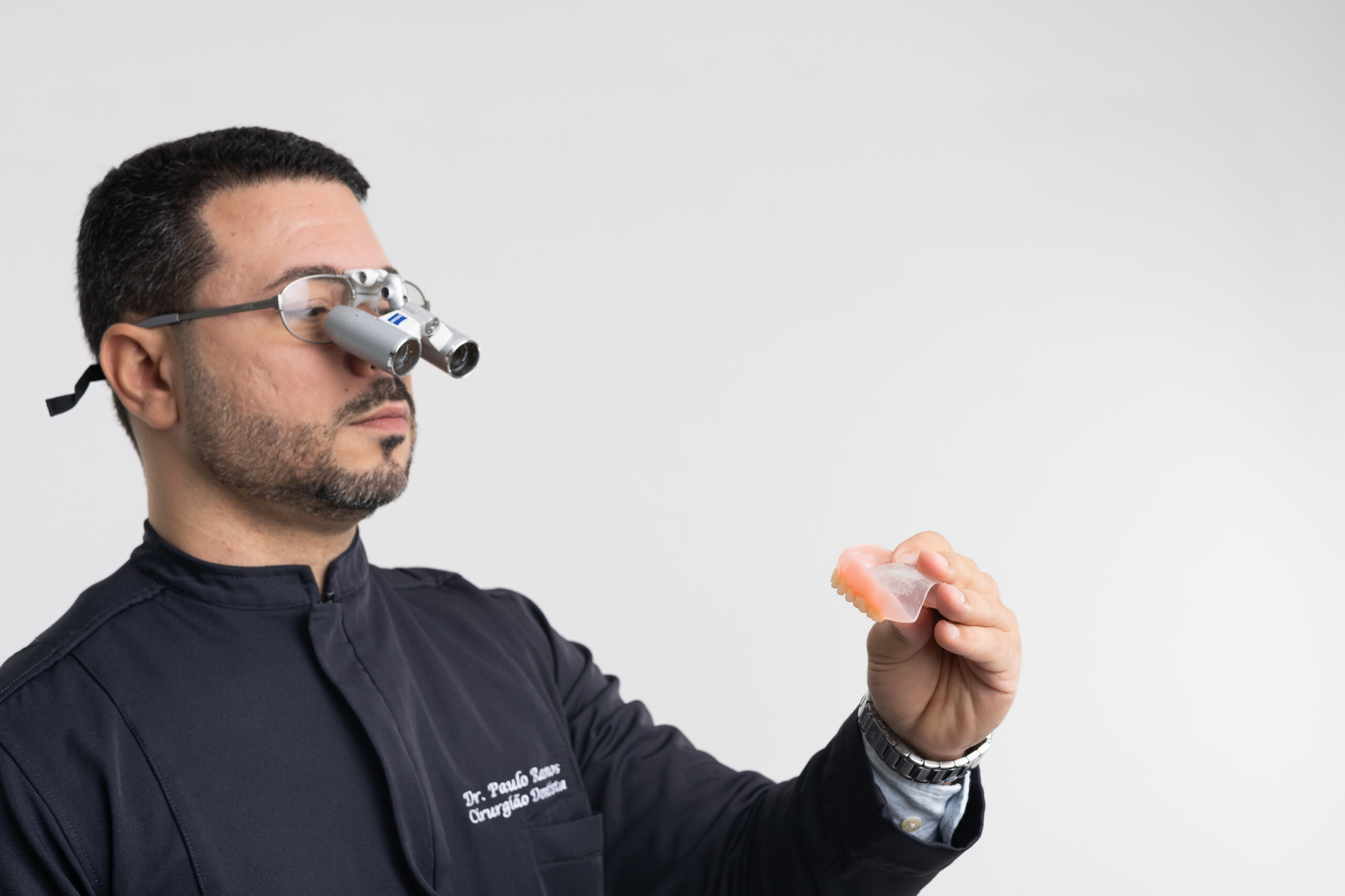 Fotografia profissional de um dentista analisando uma prótese dentaria, usando jaleco azul marinho , em frente a um fundo branco. Ele está segurando uma prótese dentaria, demonstrando confiança e profissionalismo. No jaleco, está bordado o nome “Dr. Paulo Ramos – Cirurgião-Dentista”. A imagem transmite profissionalismo e acolhimento, ideal para sites e materiais de clínicas odontológicas.
