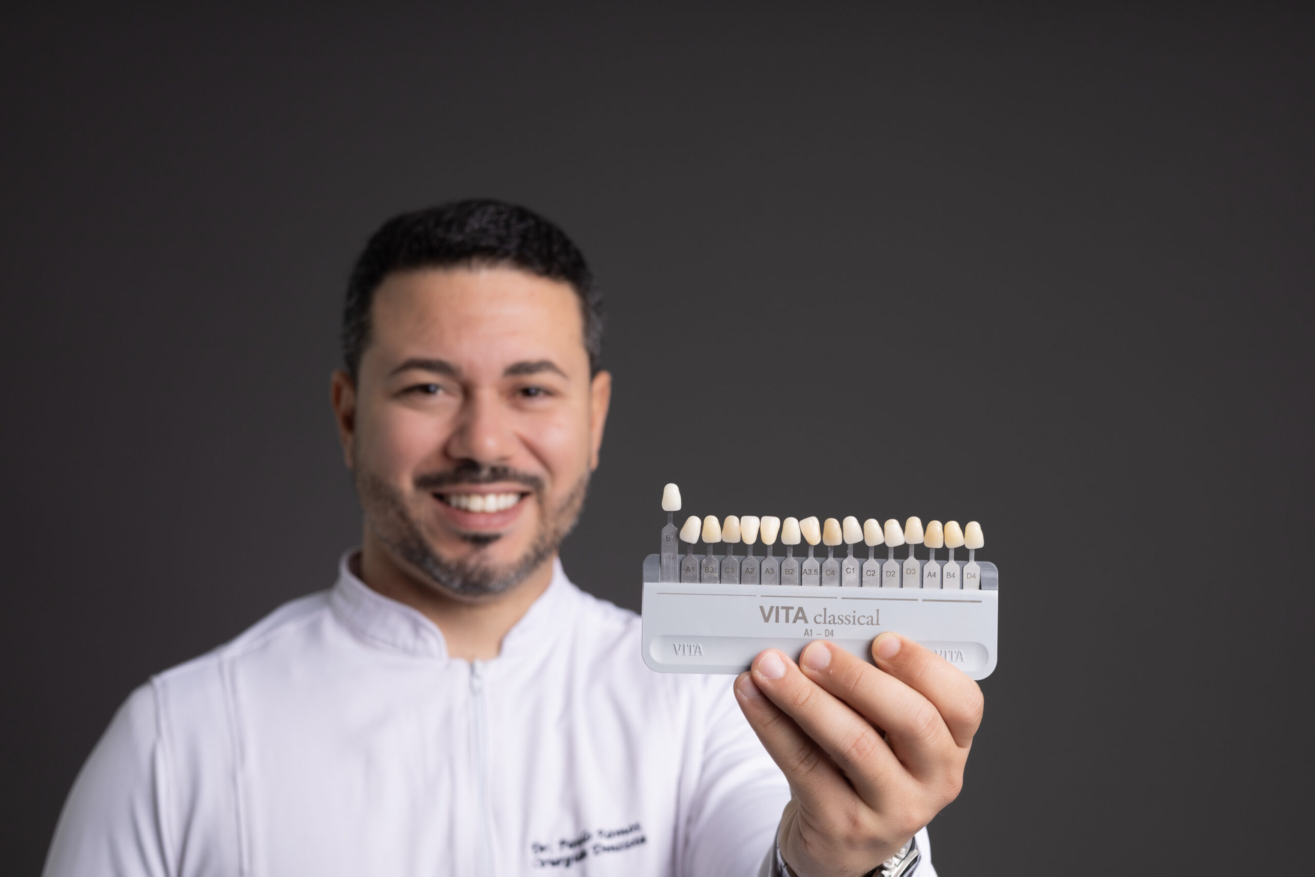 Fotografia profissional de um dentista sorridente usando jaleco branco, em frente a um fundo cinza escuro. Ele está segurando uma cartela de cor para auxiliar em lentes de contato, facetas e clareamento dental, demonstrando confiança e simpatia. No jaleco, está bordado o nome “Dr. Paulo Ramos – Cirurgião-Dentista”. A imagem transmite profissionalismo e acolhimento, ideal para sites e materiais de clínicas odontológicas.